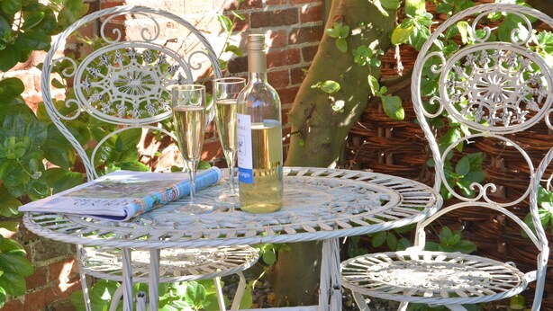 A white metal table with a bottle and glasses, surrounded by greenery. - Pebble Beach Cottage, Aldeburgh (Aldeburgh)