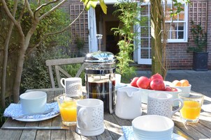 Preparación para el desayuno con cafetera, frutero y diferentes tazas y platos sobre una mesa al aire libre.