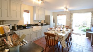 A kitchen with white cabinets, a dining table set for four, and a wicker chair. - Pebble Beach Cottage, Aldeburgh (Aldeburgh)