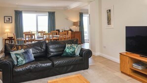 A living room with a black leather sofa, wooden coffee table, and a TV stand. There is a dining area with wooden chairs and a table in the background. - Seadrift (Saxmundham)