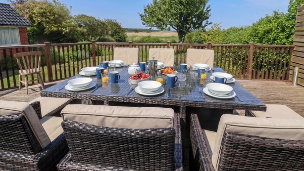 An outdoor dining area with a table set for four, surrounded by wicker chairs with cushions. - Seadrift (Saxmundham)