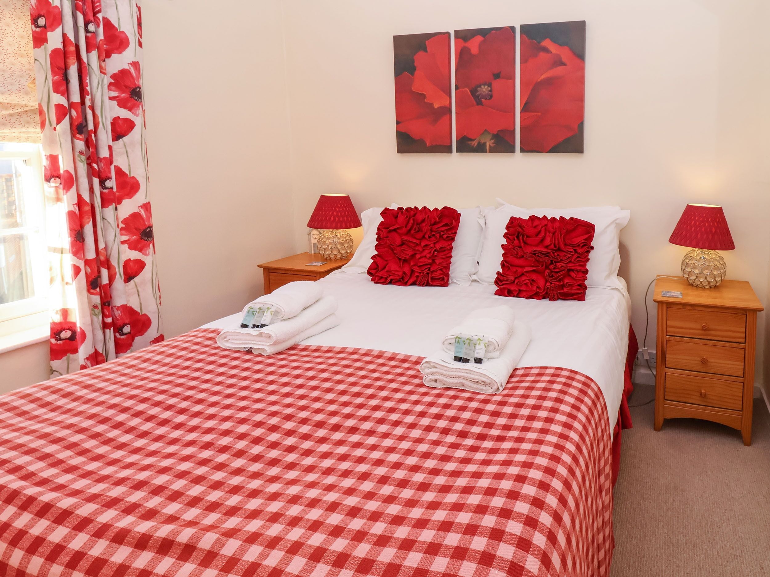 A neatly made bed with a red and white checkered bedspread, two red pillows, and two bedside tables with lamps.