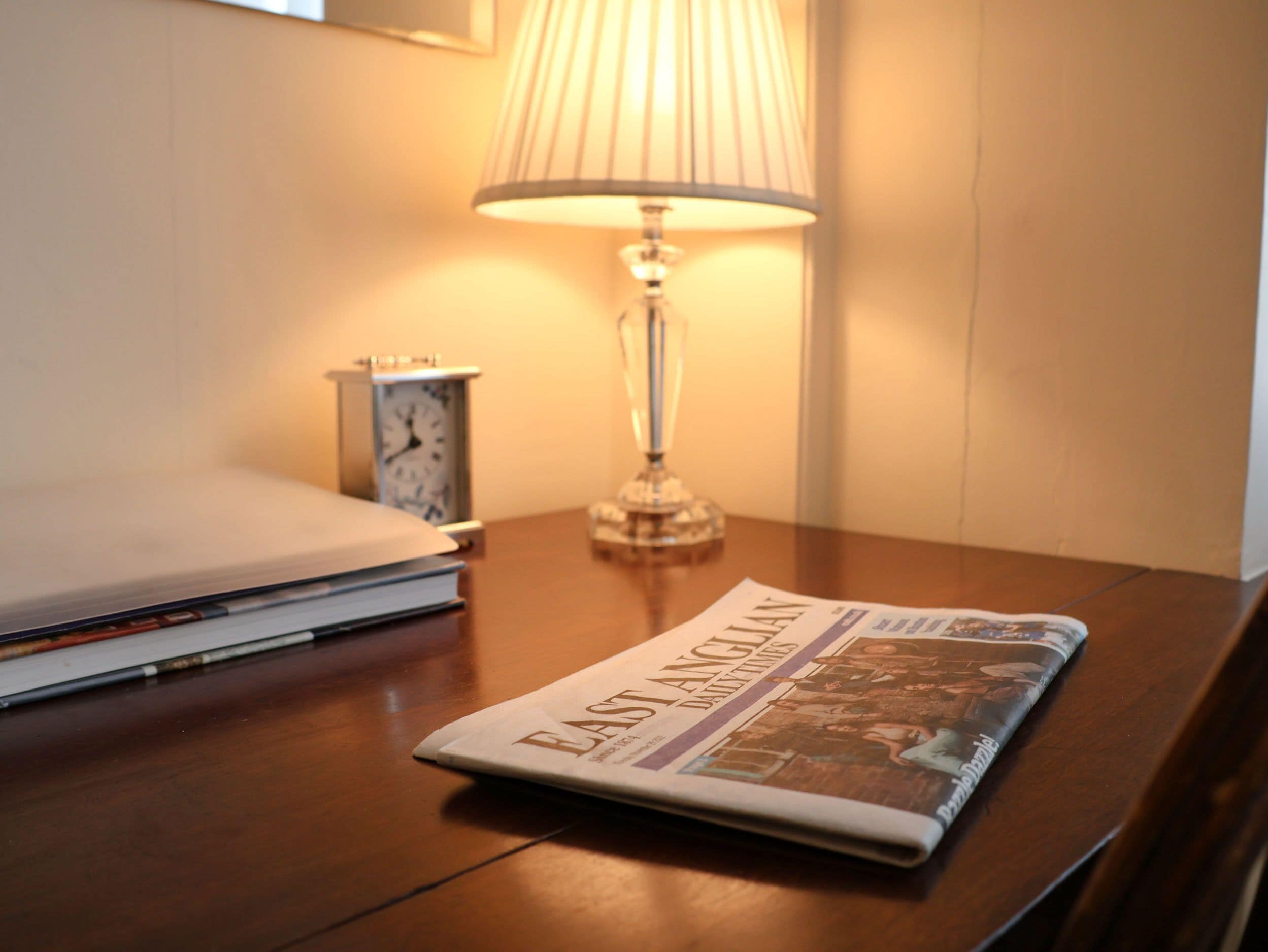 A neatly made bed with a lamp and a clock on a bedside table, and a folded newspaper on the table.