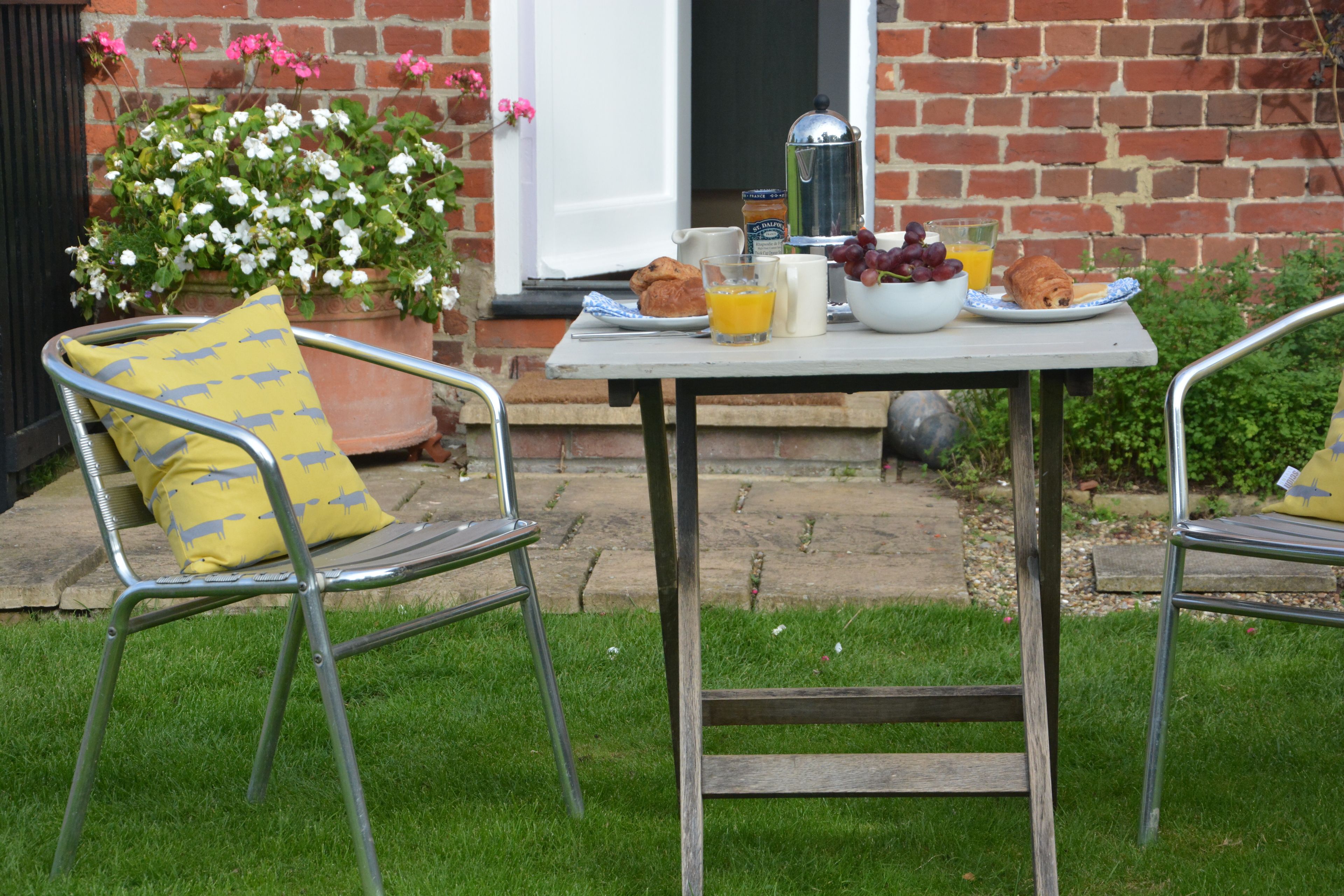 A patio with a table set for breakfast, including a coffee pot, croissants, grapes, orange juice, and a bowl of fruit.