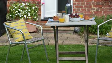 A patio with a table set for breakfast, including a coffee pot, croissants, grapes, orange juice, and a bowl of fruit.