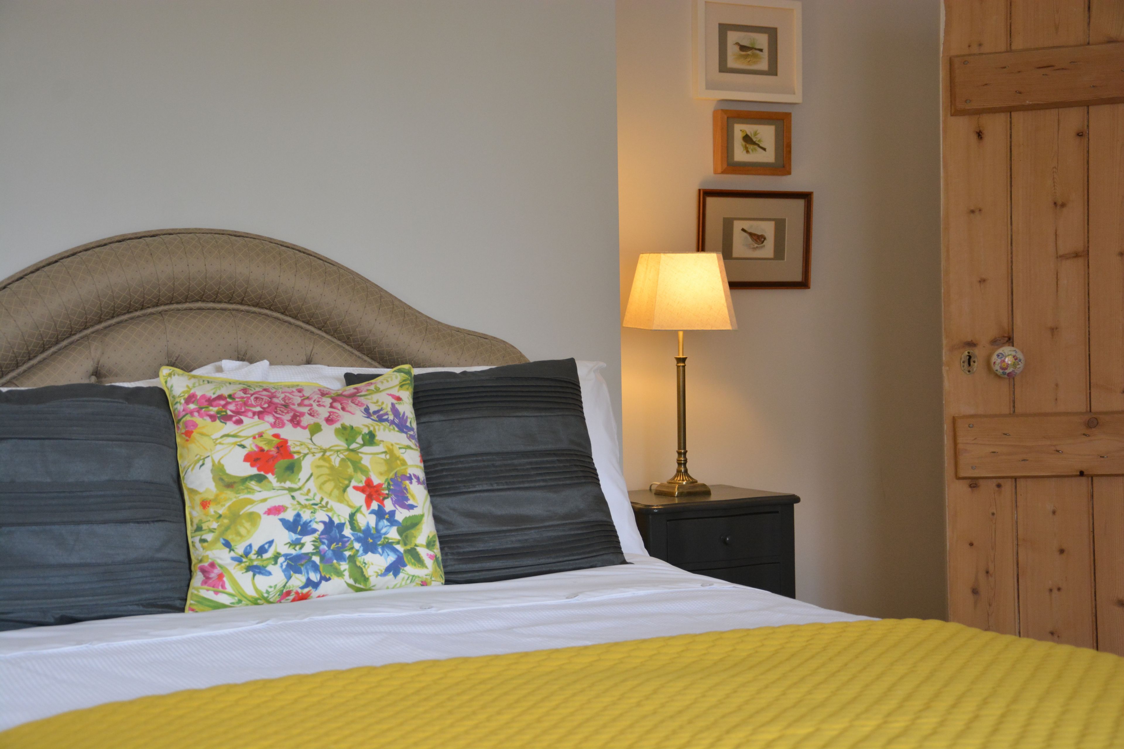 Un lit bien fait, agrémenté d’un coussin à motifs floraux, une lampe de chevet et une porte en bois en arrière-plan.