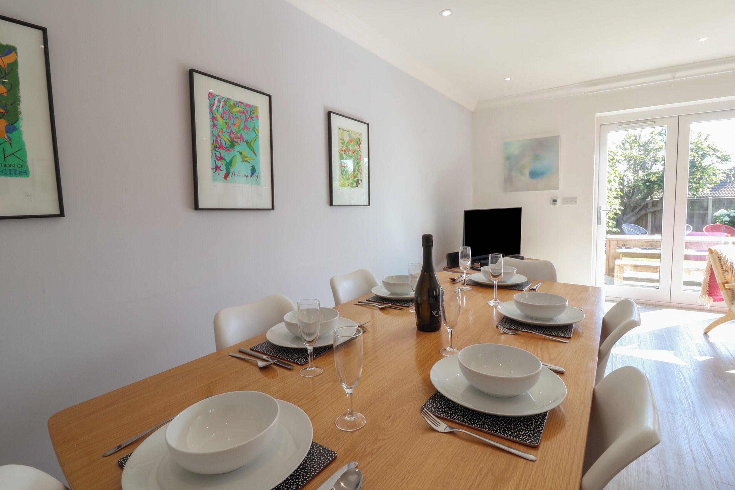 Un coin repas avec une table en bois dressée pour quatre personnes, des chaises blanches et des œuvres d’art encadrées accrochées aux murs.