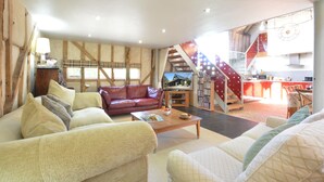 A spacious living room with a red leather sofa, a beige sofa, and a wooden coffee table. There is a television on a stand, a bookshelf, and a staircase leading to an upper level.