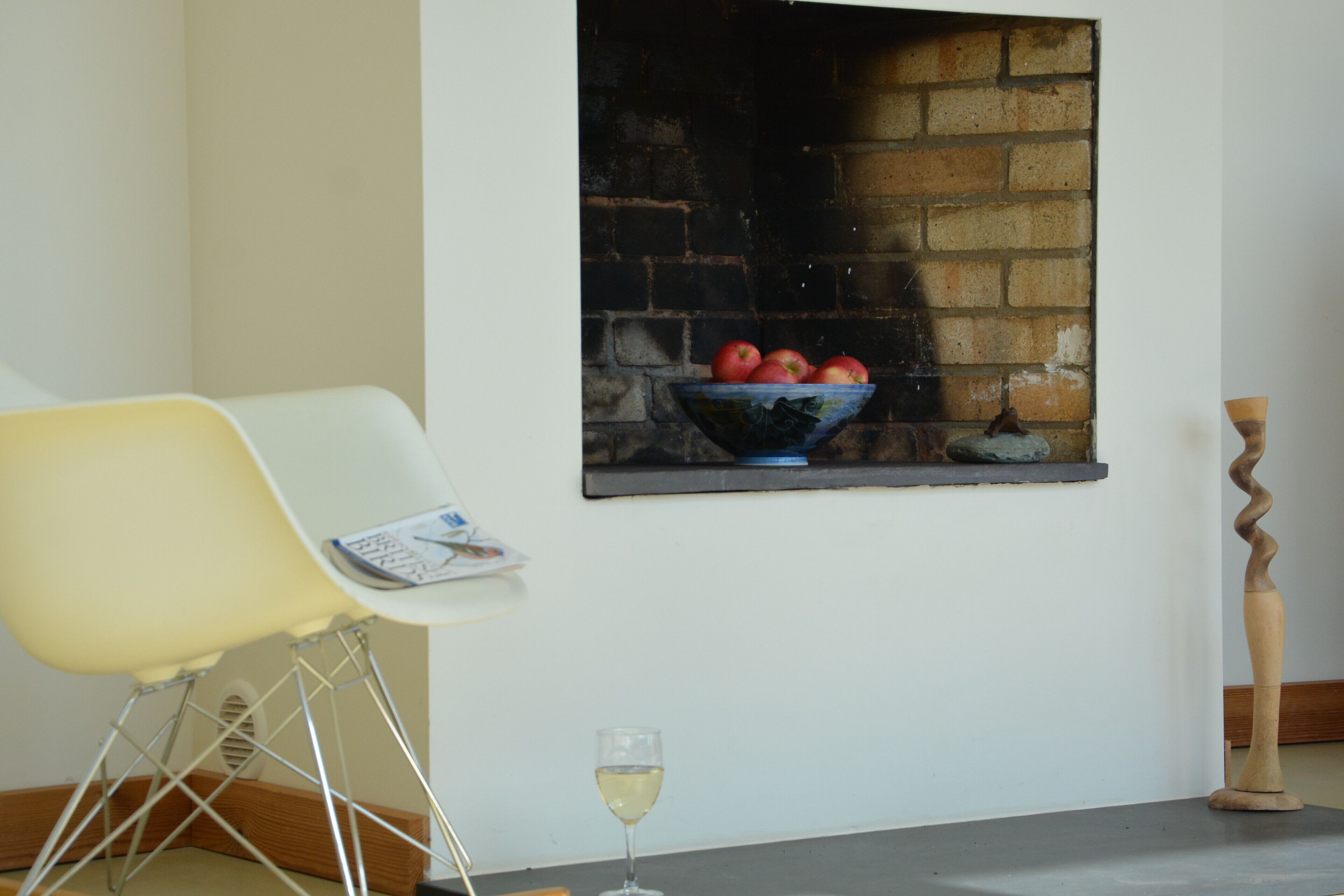 A modern living room with a white chair, a bowl of apples on a brick wall, a glass of wine, and a wooden sculpture.