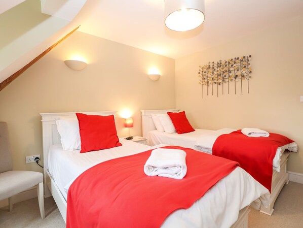 A hotel room with two beds, each with white bedding and red pillows. There is a bench, a wall-mounted light fixture, and a decorative wall piece. - Saffron, Blythview (Halesworth)