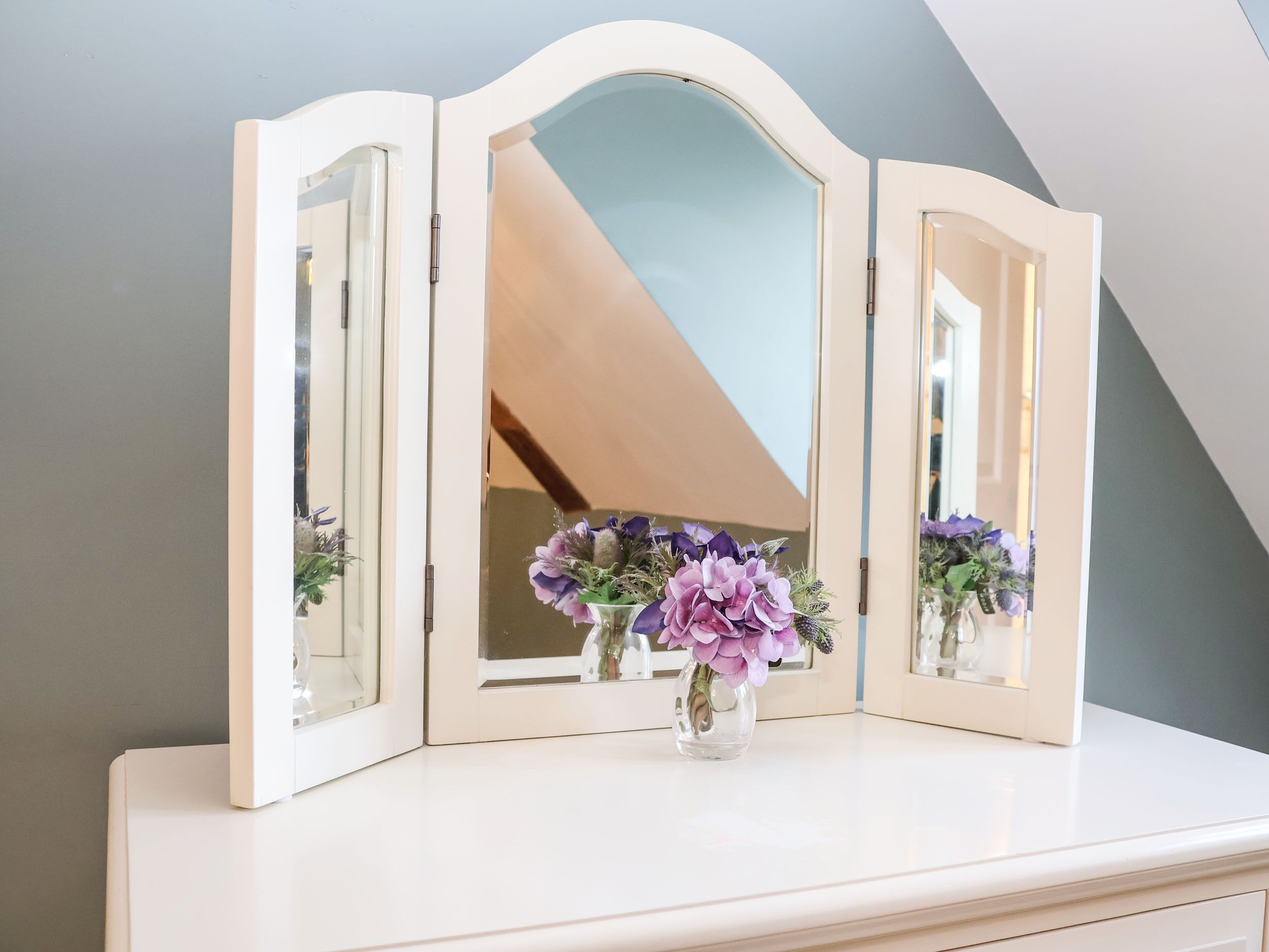 Une coiffeuse blanche avec un miroir et un vase de fleurs.