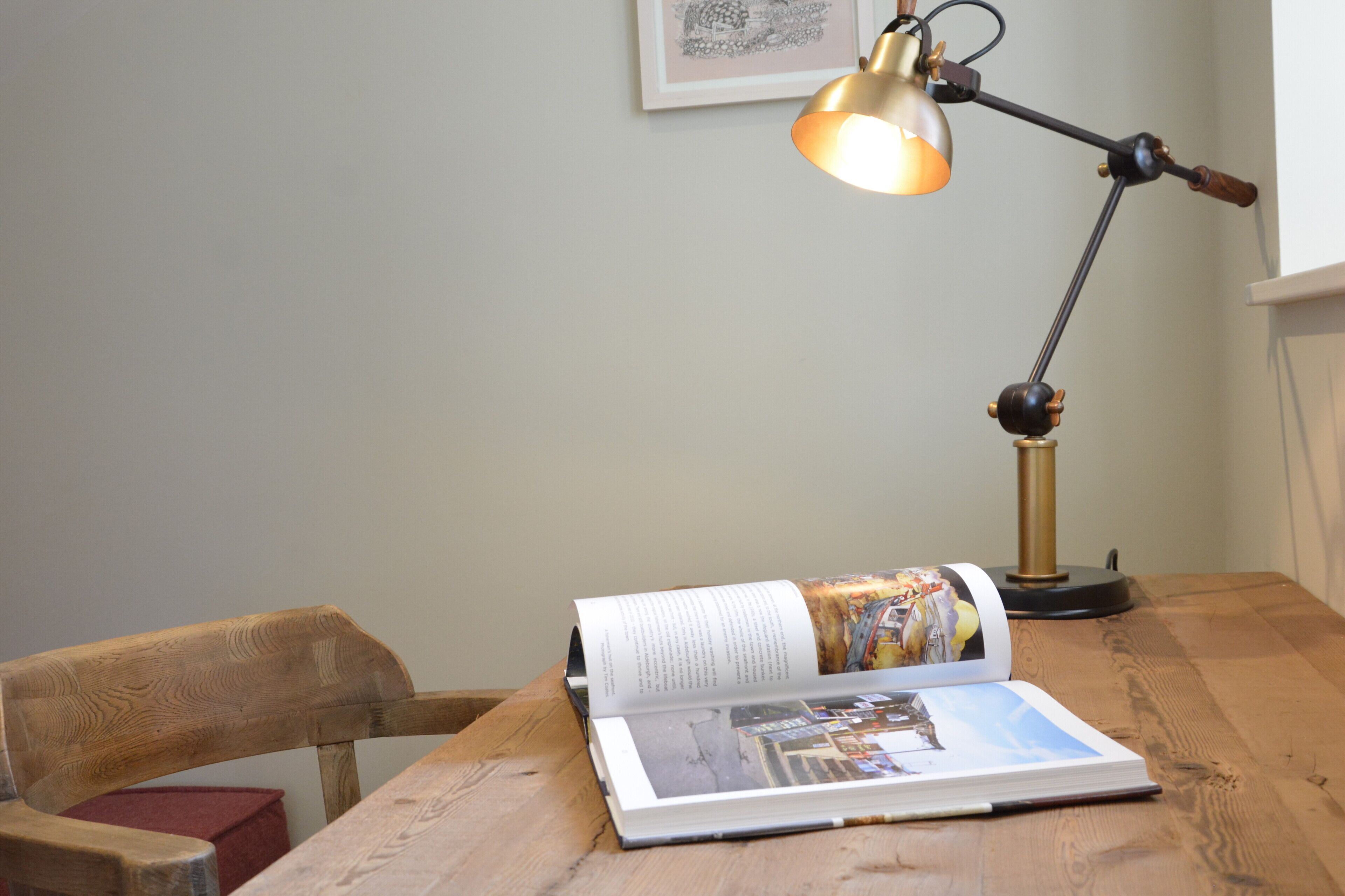 A wooden table with a book open, a desk lamp, and a framed picture on the wall.