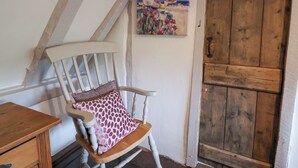 A wooden rocking chair with a patterned cushion, a wooden table, and a rustic wooden door. - Pudding Cottage, Westleton (Saxmundham)