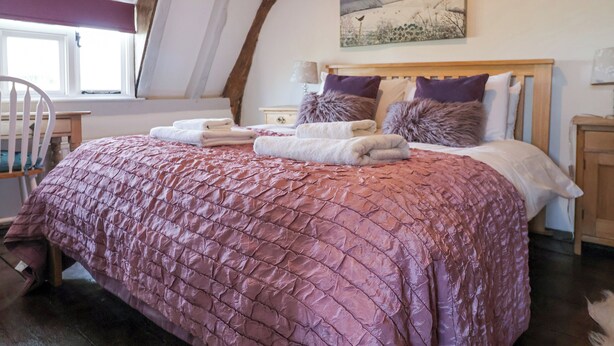 A bedroom with a wooden ceiling, a bed with a pink quilt, and a painting above the bed. - Pudding Cottage, Westleton (Saxmundham)