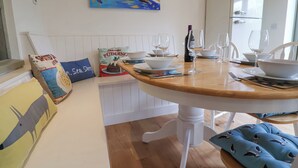 A dining area with a wooden table set for two, white chairs, a painting above the table, and a white refrigerator in the background. - Pudding Cottage, Westleton (Saxmundham)