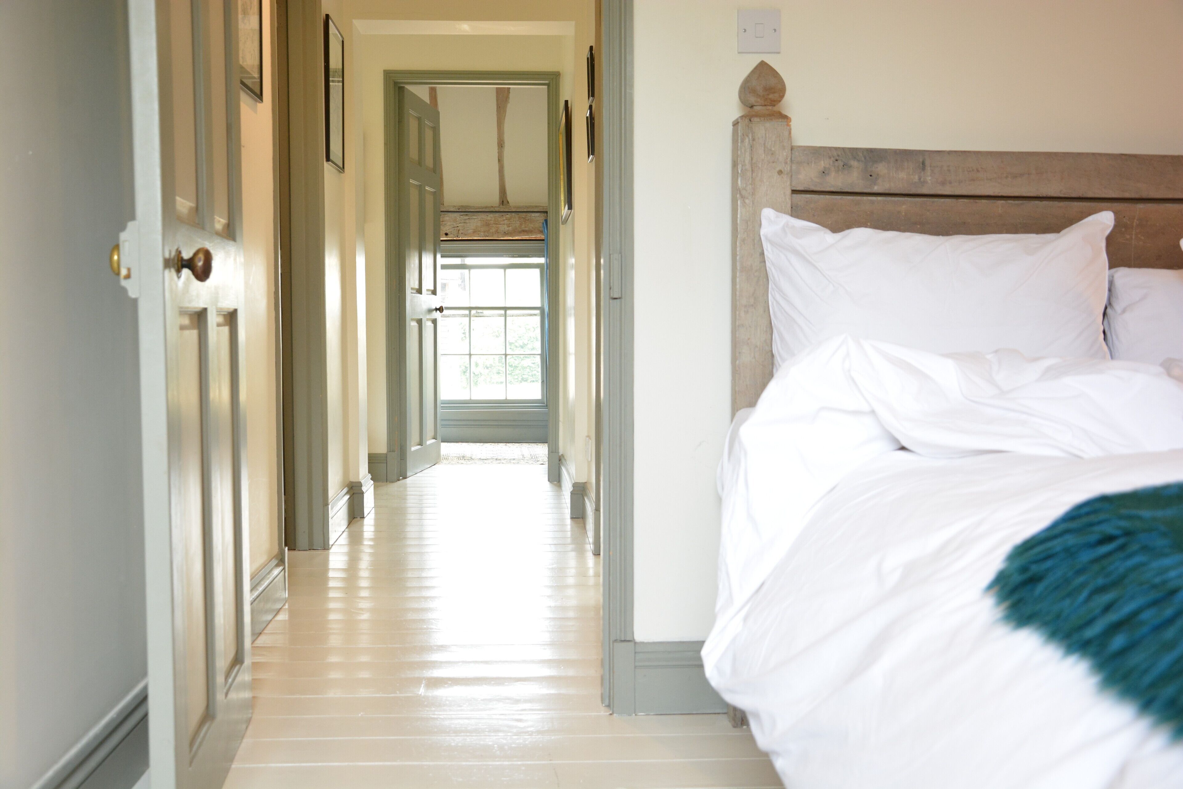 A hotel room with a wooden headboard, a bed with white linens, and a hallway leading to a bright room with a window.