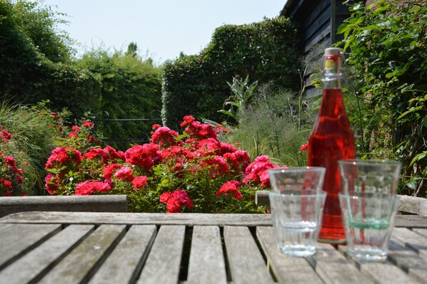 A wooden table with two clear glasses and a red bottle, surrounded by vibrant red flowers and greenery. - The Gig House, Stowupland (Stowmarket)