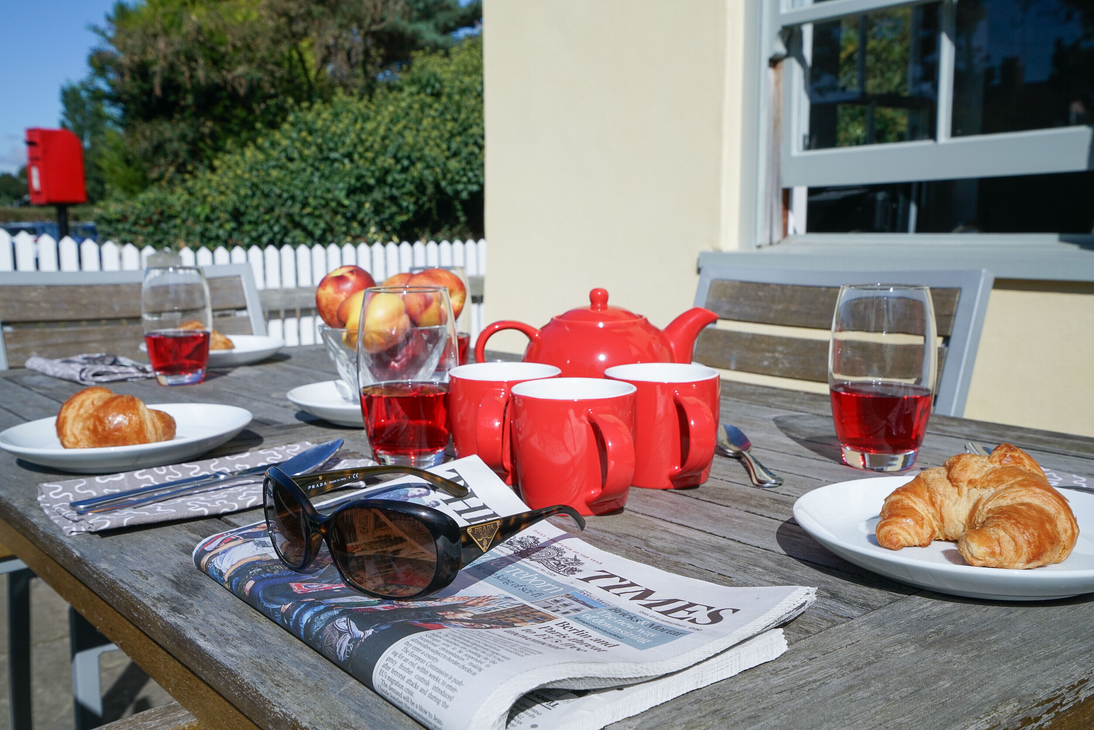 Ein Picknick mit Zeitung, Croissants, Äpfeln, einem Teekessel und roten Tassen auf einem Holztisch.