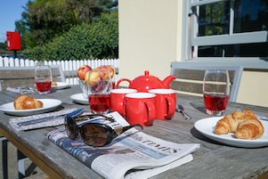 A picnic setup with a newspaper, croissants, apples, a teapot, and red cups on a wooden table.