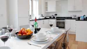 A modern kitchen with white cabinets, a dining table set with bowls, plates, and wine glasses, and a bowl of fruit on the table. - Seal Cove (Reydon)