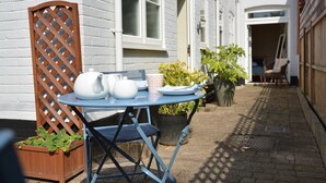 A small outdoor table with a blue tablecloth, a white teapot, a white jug, and a patterned cup. There are potted plants and a wooden chair in the background. - Spindrift, Southwold (Reydon)