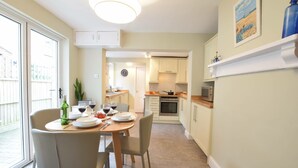 A dining area with a table set for four, a clock on the wall, and a view of the kitchen and balcony. - Spindrift, Southwold (Reydon)