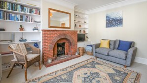 A living room with a fireplace, a bookshelf, a grey sofa with blue and yellow cushions, and a patterned rug. - Spindrift, Southwold (Reydon)