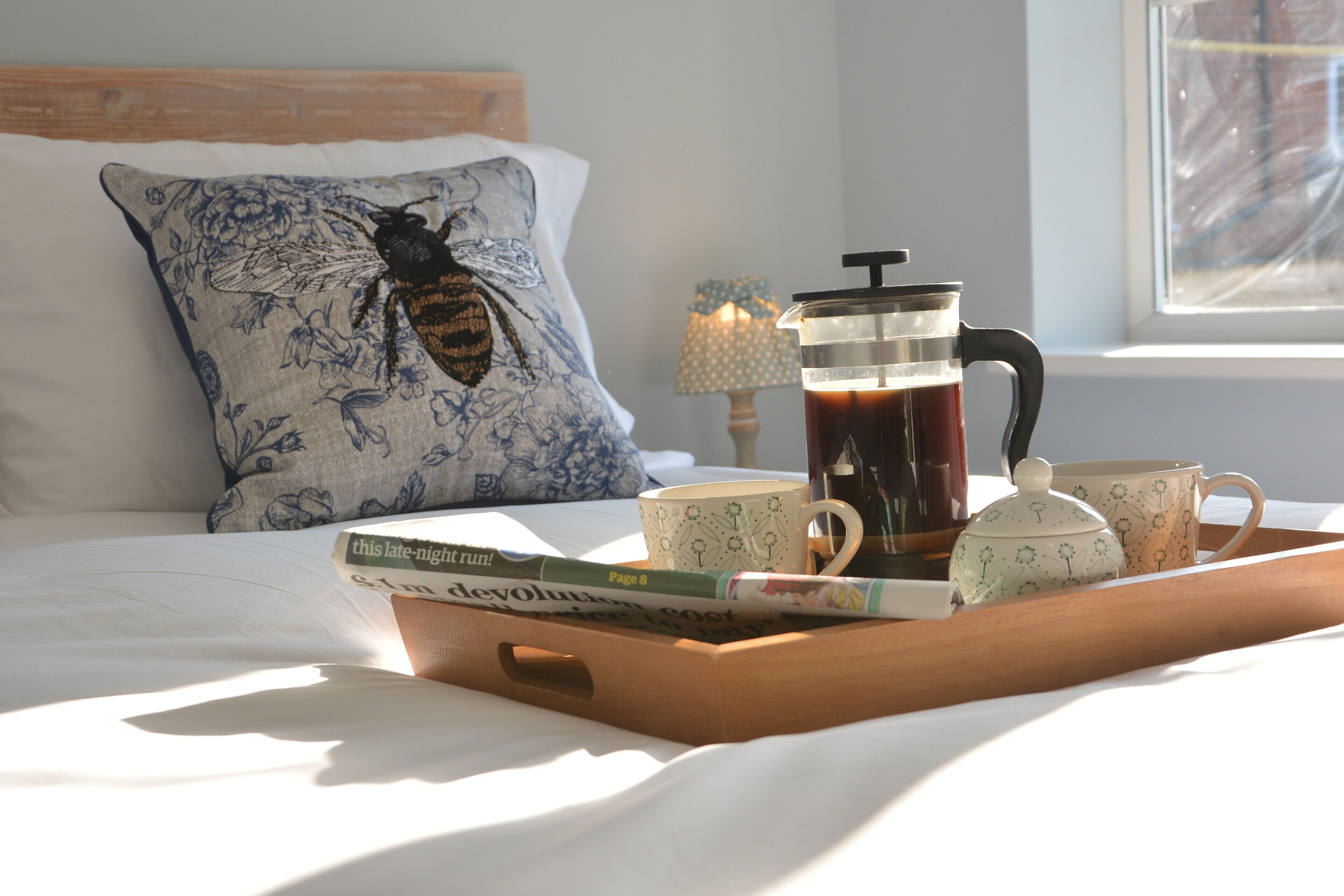 A wooden tray with a coffee pot, teapot, and cups on a bed with a floral pillow and books.