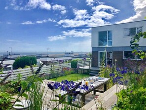 A modern building with a large window, a patio with a table set for a meal, and a view of the sea. - Aldeburgh Lodge Gardens (Aldeburgh)
