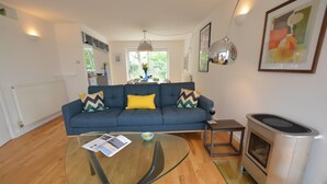 A modern living room with a blue sofa, a glass coffee table, a wood-burning stove, and a dining area in the background.