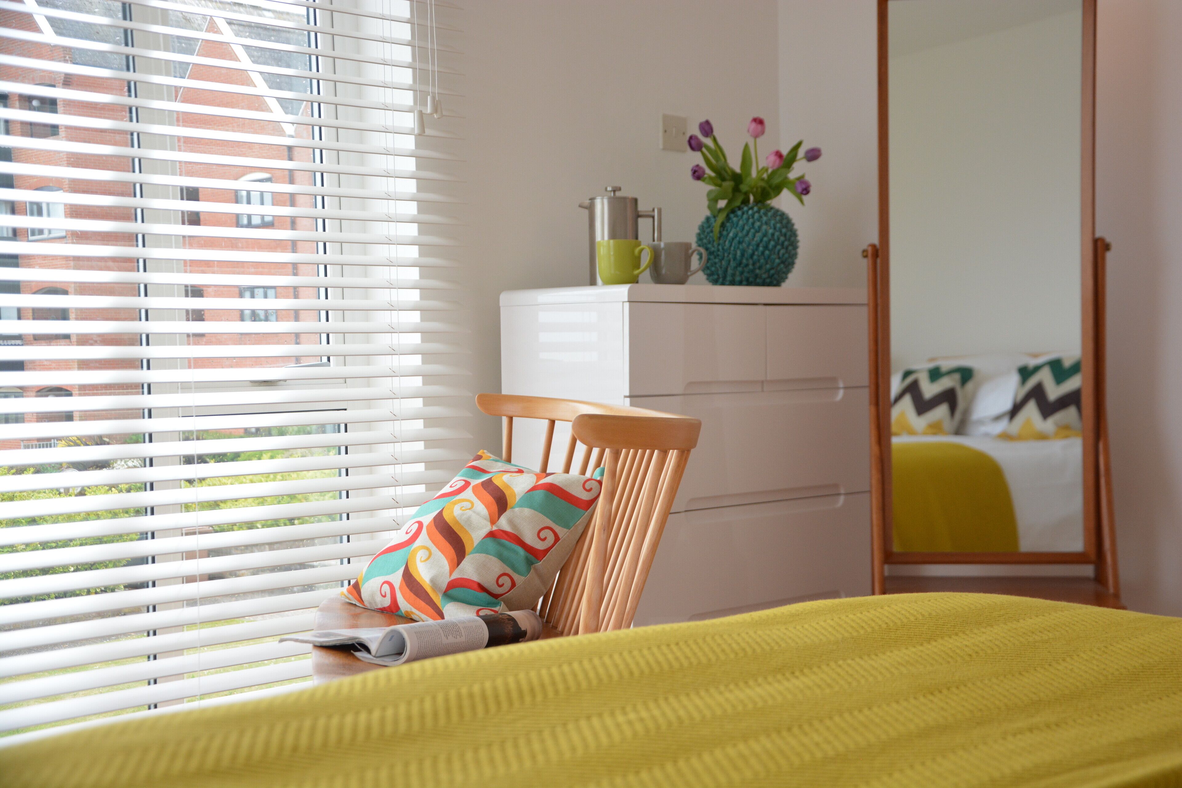 A bedroom with a bed, a chair, a dresser, a mirror, and a window with blinds.