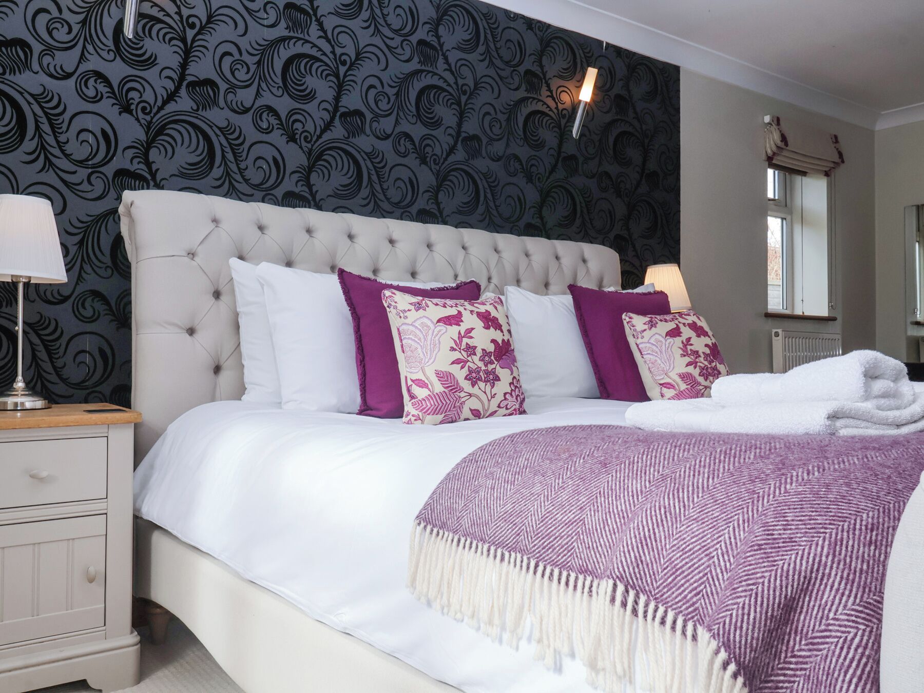 A bedroom with a tufted headboard, a bed with white and purple bedding, a bedside table with a lamp, and a wall with a black floral wallpaper.