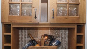 Un meuble en bois doté de portes en verre, dans lequel sont rangés des verres à vin. En dessous, une boîte en bois contient une bouteille de champagne, un sachet de poulet rôti aux herbes et un tissu à carreaux.