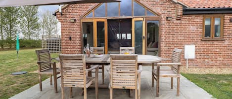 A brick building with a patio area featuring a table and chairs, a large umbrella, and a view of the interior through glass doors.