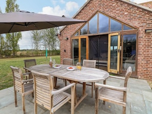 Une terrasse extérieure avec une table et des chaises en bois, une porte vitrée donnant sur un espace abrité et un bâtiment en briques en arrière-plan.