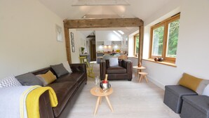 A modern living room with a brown leather sofa, a grey sofa, and a wooden coffee table with a bottle of champagne. There is a kitchen visible in the background. - Red Poll Barn (Halesworth)