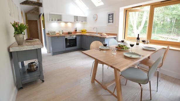 A modern kitchen with a wooden dining table, grey cabinets, and a window with a view of trees. - Red Poll Barn (Halesworth)