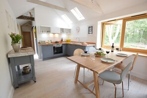 A modern kitchen with a wooden dining table, grey cabinets, and a window with a view of trees. - Red Poll Barn (Halesworth)