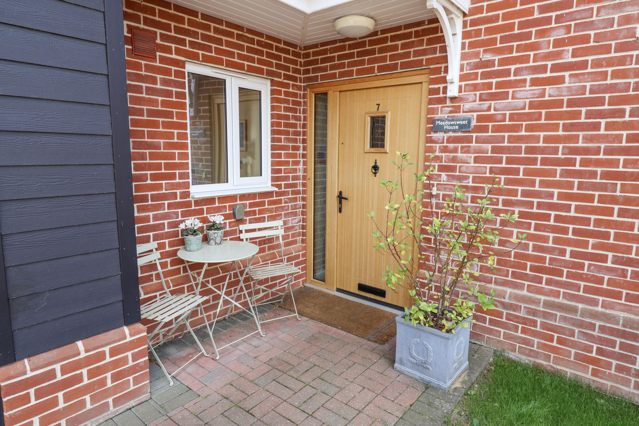 A brick house with a wooden door, a small garden area with a table and chairs, and a potted plant.