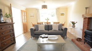 A living room with a sofa, coffee table, and fireplace. There is a dining table with chairs and a window with curtains. - Meadowsweet, Blythburgh (Halesworth)