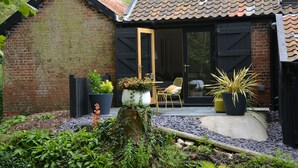 A brick building with a tiled roof, a wooden door, and a small patio area with potted plants and a chair. - Milk Parlour (Halesworth)
