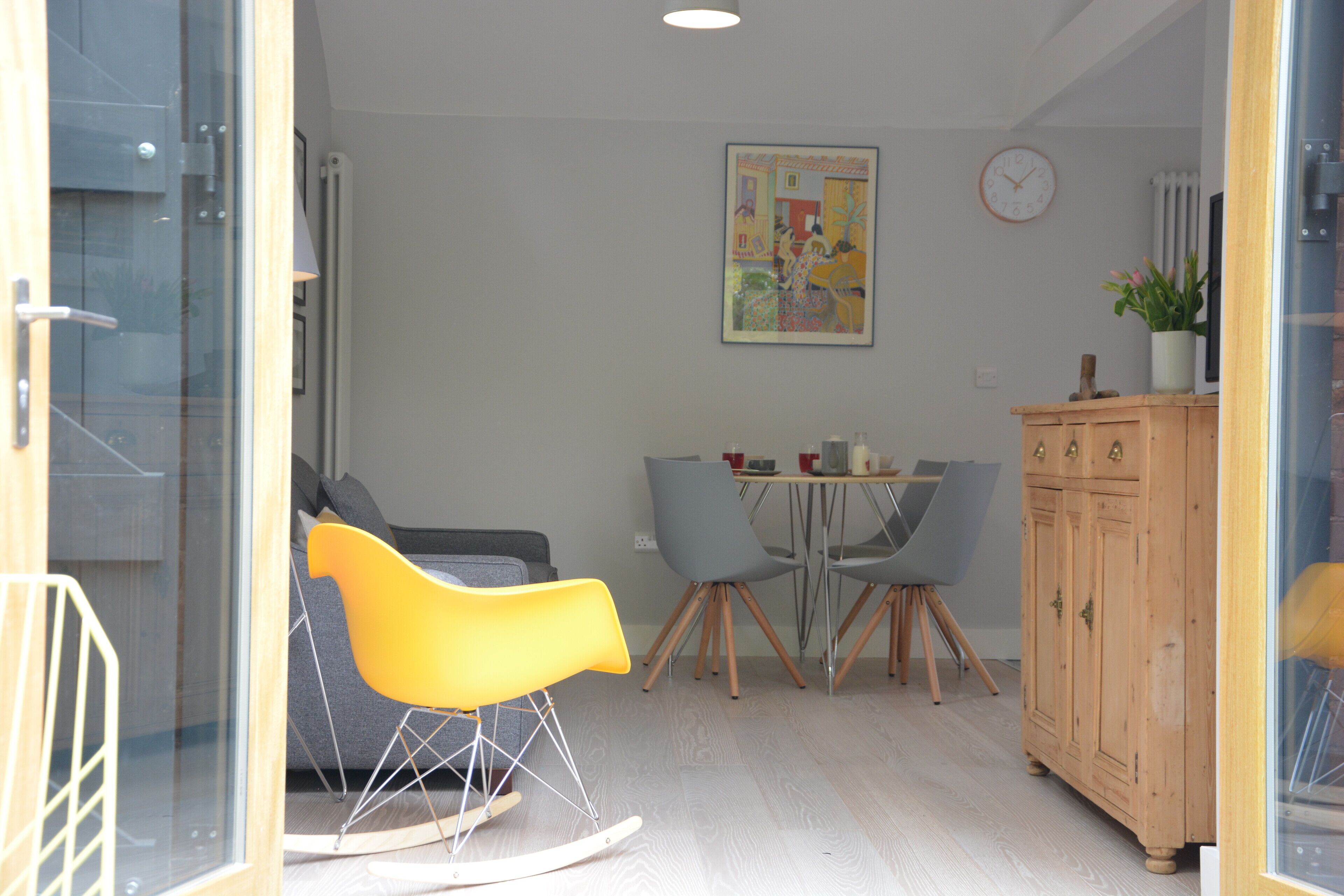 Een moderne woonkamer met een eetgedeelte, met een houten kast, een eettafel met stoelen en een kleurrijk abstract schilderij aan de muur.