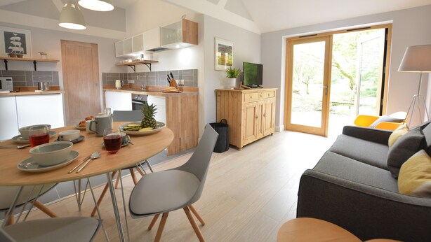 A modern kitchen with a dining area, a grey sofa, and a TV on a wooden cabinet. - Milk Parlour (Halesworth)