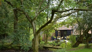 A rustic house with a brick exterior and a tiled roof, surrounded by lush greenery and a small wooden bridge over a pond. - Milk Parlour (Halesworth)