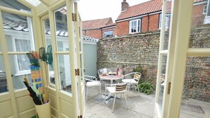 A conservatory with a stone wall, a table with chairs, and a door leading to a room with a patterned rug and a bucket. - Walton House, Southwold (Reydon)