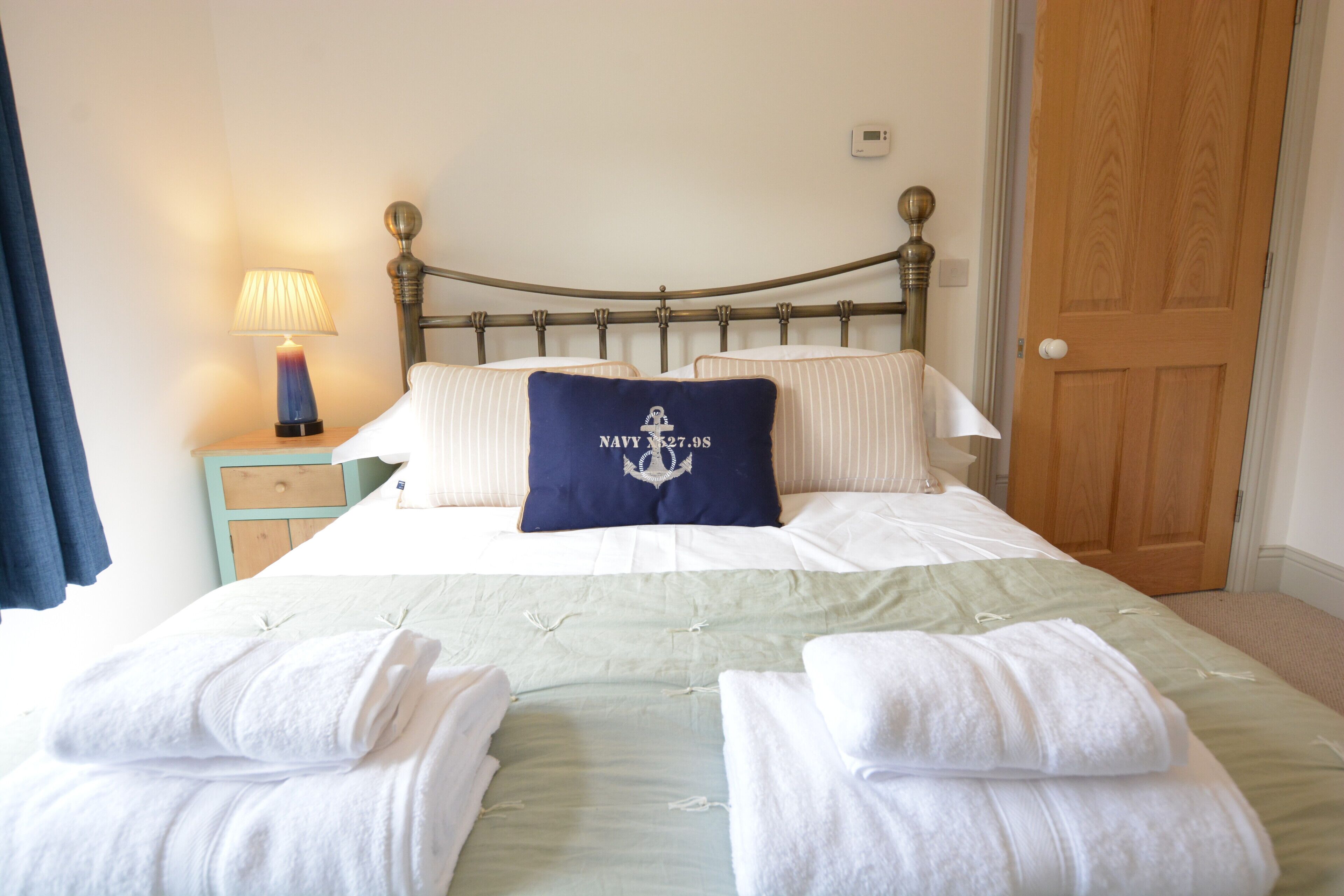Una cama bien hecha con un cojín azul marino con diseño de ancla náutica, una lámpara de noche y una puerta de madera al fondo.