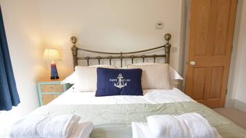 Una cama bien hecha con un cojín azul marino con diseño de ancla náutica, una lámpara de noche y una puerta de madera al fondo.