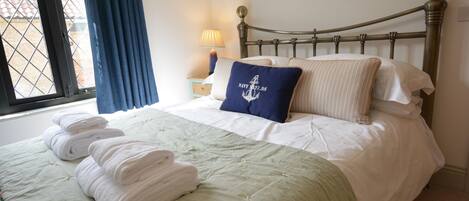 Un lit bien fait, avec une couette verte, des oreillers blancs et un coussin bleu marine orné d’un motif nautique. Des serviettes blanches pliées sont posées sur le lit, et une lampe se trouve sur la table de chevet, à côté du lit.