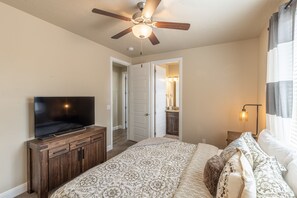 A bedroom with a bed, a TV on a wooden cabinet, a ceiling fan, and a bathroom visible through an open door.