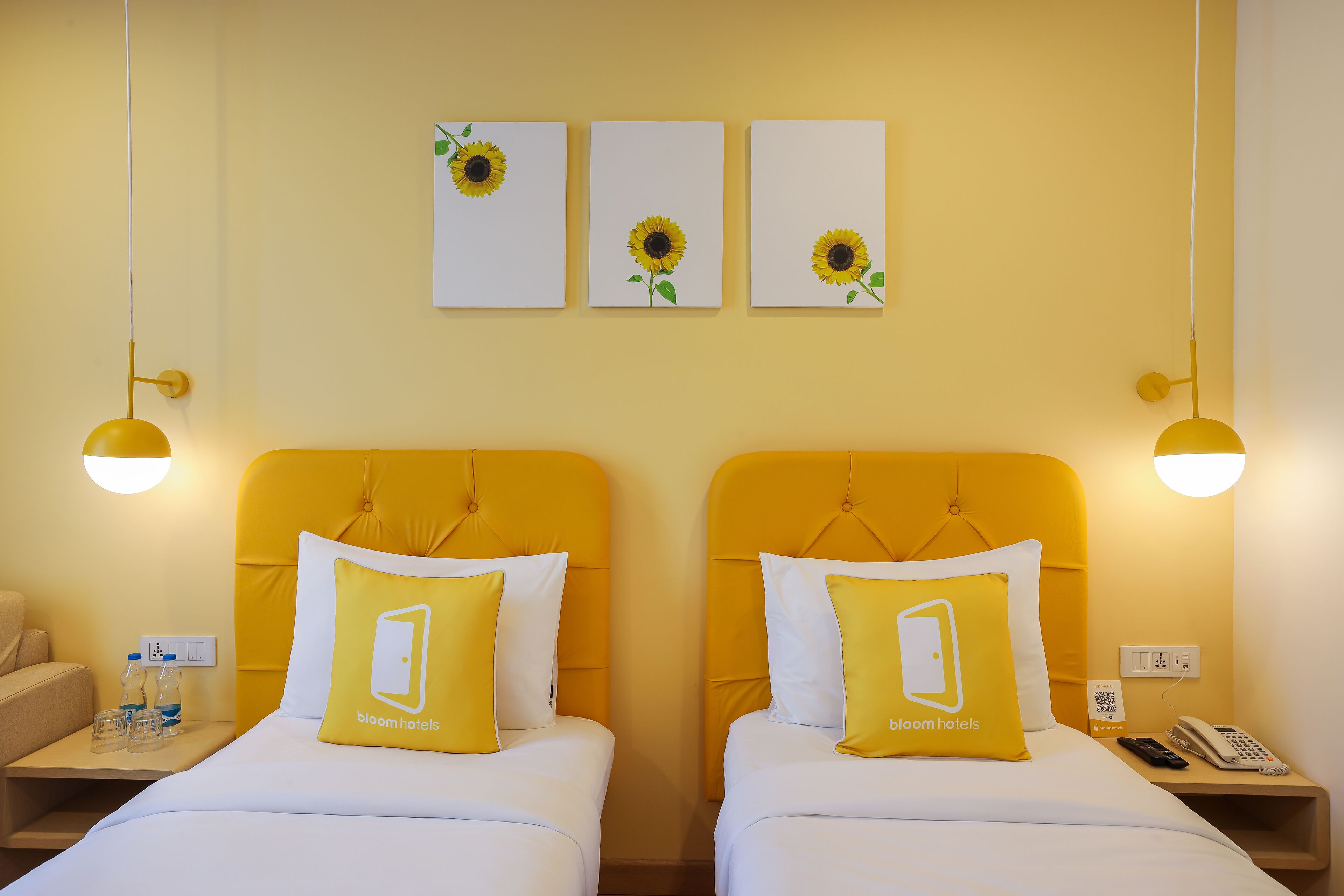 A hotel room with two beds, each with a yellow headboard and a 'bloom hotels' pillow. There are three framed sunflowers on the wall, and the room has a yellow and white color scheme.